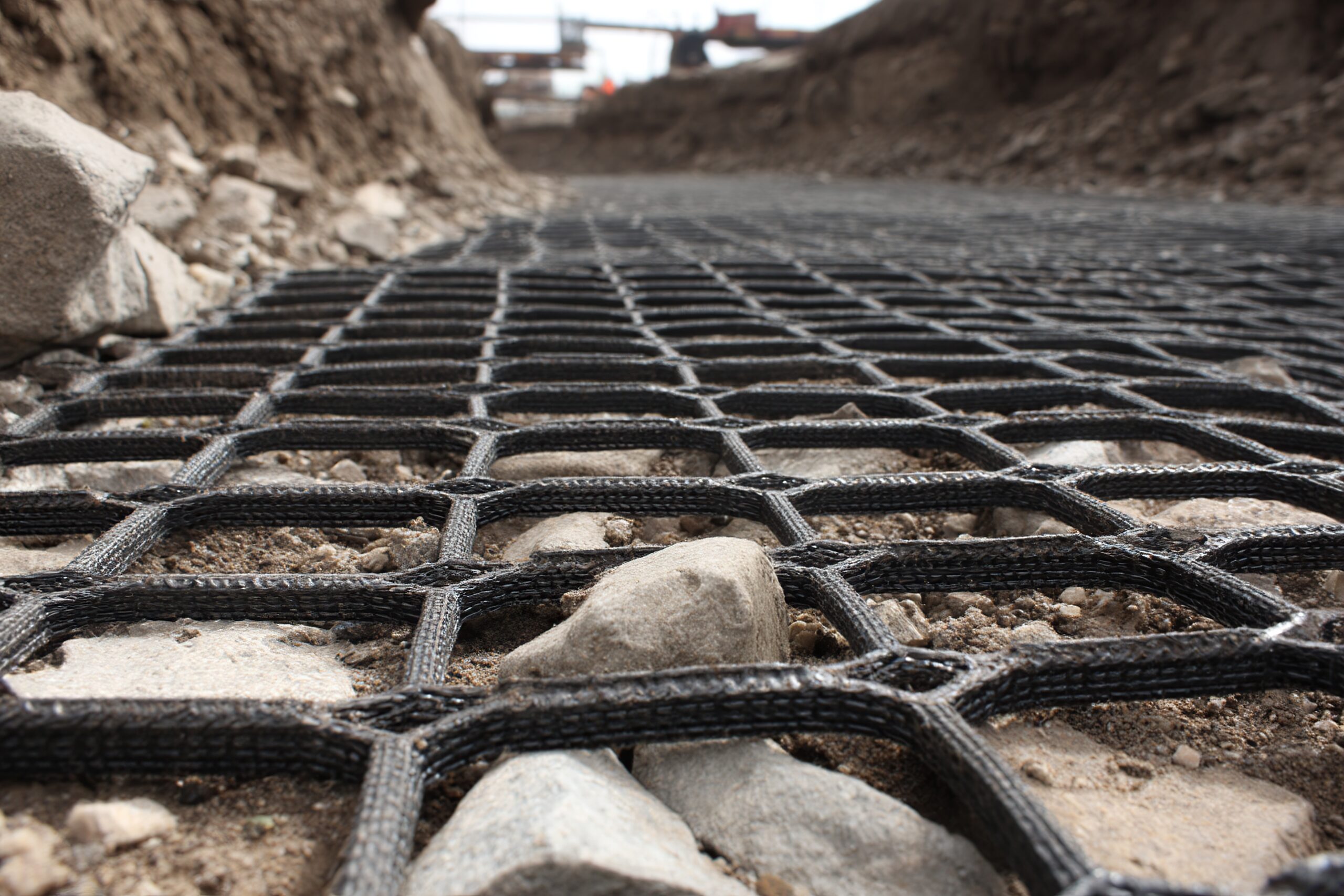 Road Reinforcement Grid with Construction Site | Reedy Chemical Foam Close-up of a grid of steel rebar laid out on rocky ground at a construction site, with dirt and piles of soil on either side and a blurred background indicating ongoing work.