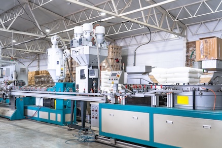 Plastic Extrusion Manufacturing Machine in Modern Industrial Factory Automated Polymer Processing Line for Plastic Products Production with Raw Material and Control Panels | Reedy Chemical Foam Industrial plastic extrusion line in a bright factory, with white hoppers feeding material into a long blue machine.