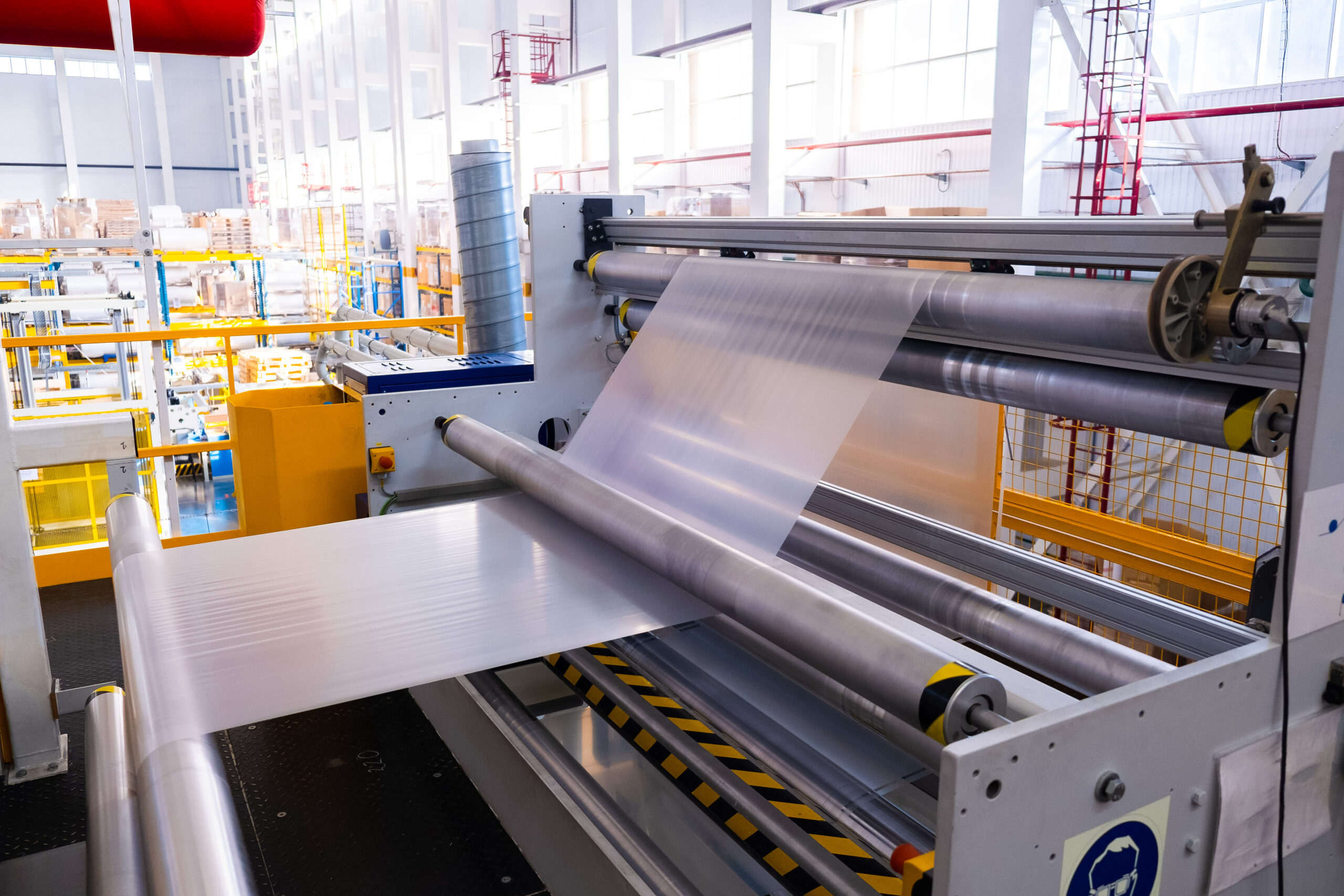 Industrial Line for the Production of Plastic Packaging Film | Reedy Chemical Foam Automated metal sheet roller system in a bright industrial workspace with yellow safety rails besides the conveyor.